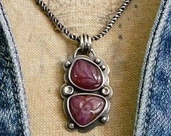 May include: A silver chain necklace with a pendant featuring two carved pink stones set in silver. The necklace is worn on a denim jacket with a metal button that reads "CHAPS".