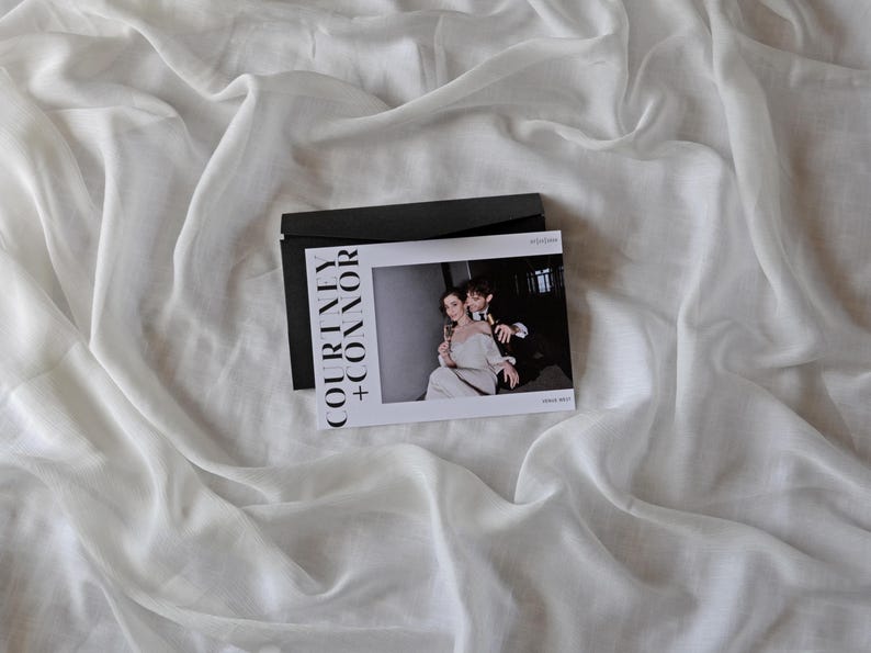 May include: A white card with black text that reads "COURTNEY + CONNOR" and a photo of a couple in a formal setting. The card is on a white fabric background.