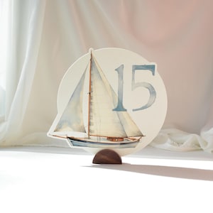 May include: A nautical-themed table number featuring a watercolor sailboat illustration. The number "15" is displayed in blue. The sign is mounted on a dark wooden base. The sailboat has white sails and a blue hull.