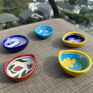 May include: Five small, hand-painted ceramic bowls in various colors. The bowls feature floral and abstract designs in blue, yellow, red, and white. The bowls are oval-shaped and sit on a textured surface.