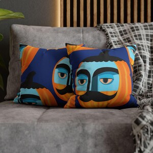 Può includere: Due cuscini decorativi con un design a volto stravagante. I cuscini presentano uno sfondo blu con tratti del viso arancioni e azzurri, inclusi i baffi. I cuscini sono su un divano grigio.