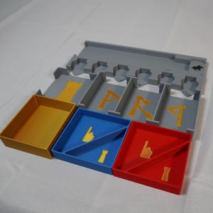 Puede incluir: Un organizador de plástico gris con compartimentos para guardar piezas de juego. Los compartimentos están etiquetados con las letras "PRA". Hay tres cajas más pequeñas, una dorada, una azul y una roja, con divisores en el interior.