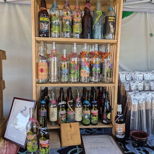 Puede incluir: Una estantería de madera con tres niveles muestra una variedad de botellas de vidrio con etiquetas coloridas. Las botellas están llenas de diferentes líquidos, incluyendo vino, ron y otras bebidas espirituosas. La estantería está instalada en un mercado o feria, con una mesa delante que muestra botellas adicionales y otros artículos.