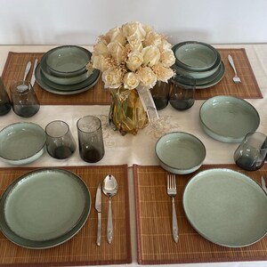 May include: A table setting with green-toned ceramic plates, bowls, and glasses. Silverware and bamboo placemats are arranged around a vase of white roses. The setting is for a formal dinner.