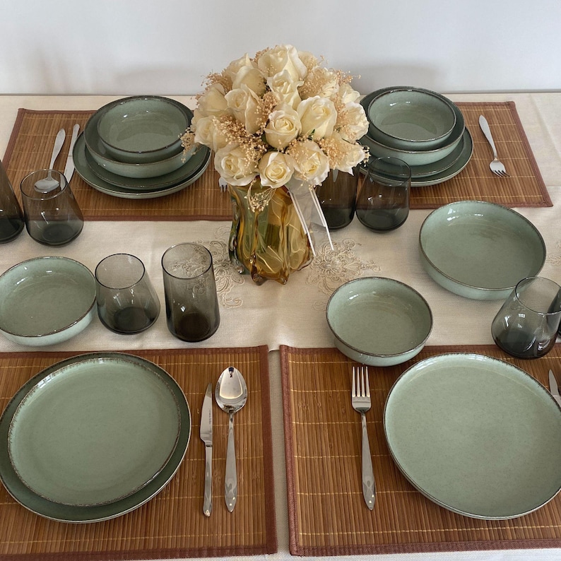 May include: A table setting with green-toned ceramic plates, bowls, and glasses. Silverware and bamboo placemats are arranged around a vase of white roses. The setting is for a formal dinner.