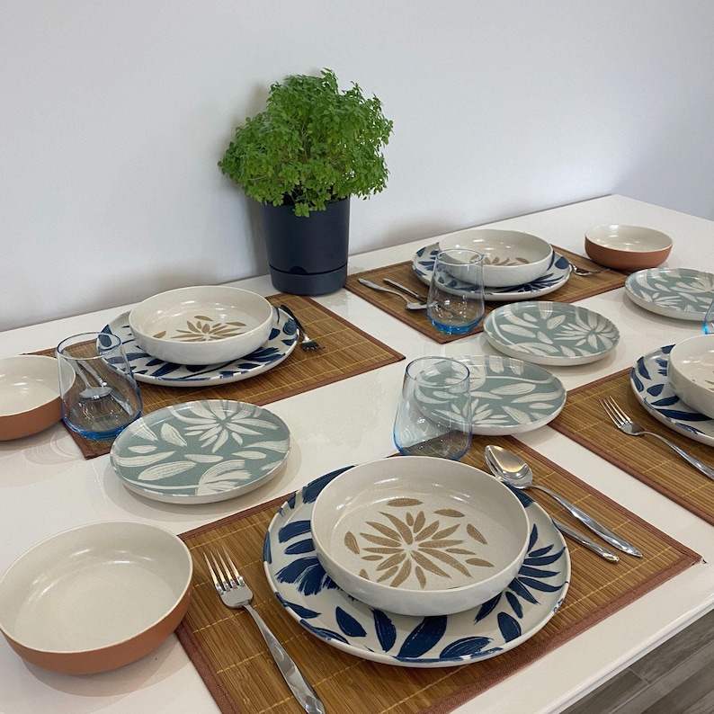 May include: A table setting with multiple place settings. Each setting includes a white bowl with a tan floral design, a matching plate with blue floral patterns, a clear glass with a blue rim, and silverware. The table also has a potted plant.