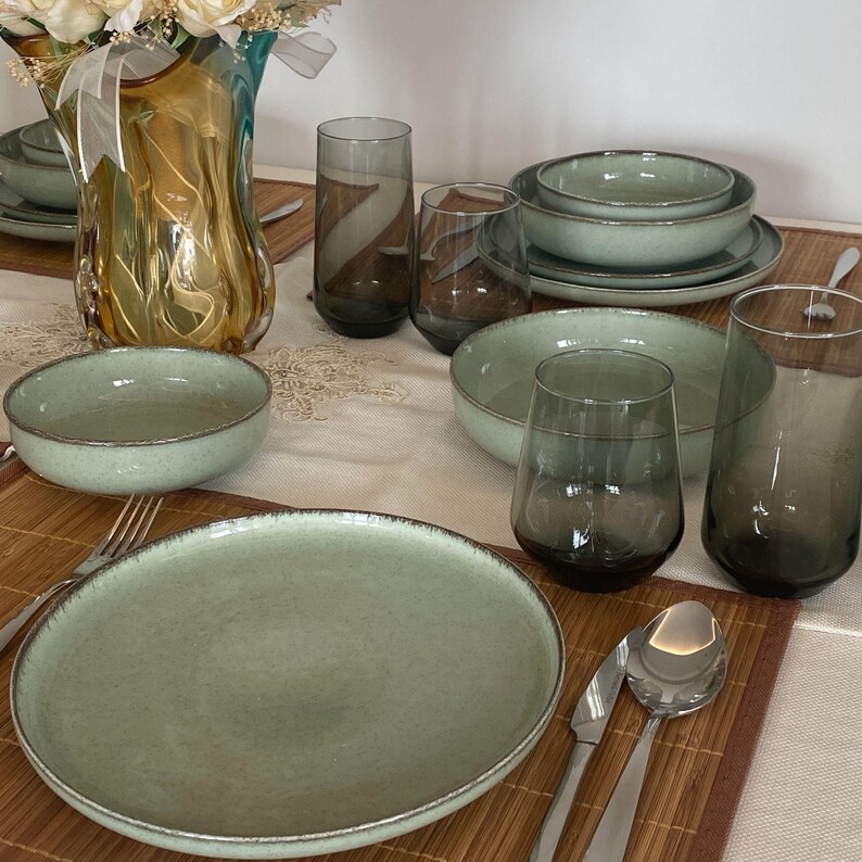 May include: A complete dinnerware set in a soft green hue, including plates, bowls, and glasses. The plates and bowls have a textured rim. Tall, smoky-gray drinking glasses and a decorative vase are also present, creating an elegant table setting.