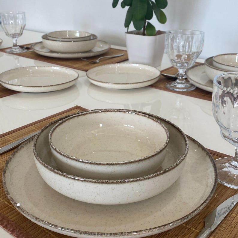 May include: A dining table setting with cream-colored ceramic plates, bowls, and glasses. The tableware has a speckled texture and a brown rim. Each place setting includes a plate, bowl, and a glass. The table is set with bamboo placemats and silverware.