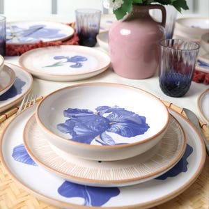 Könnte beinhalten: Ein Geschirrset aus weißer Keramik mit blauem Iris-Blumenmuster und beigen Rändern. Das Set besteht aus einer tiefen Schale, einem Salatteller mit feinen beigen Linien und einem großen Speiseteller, die alle auf einer geflochtenen Matte stehen.