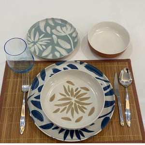 May include: A table setting featuring a blue-rimmed glass, a fork, a spoon, and a knife. The plates and bowls have floral patterns in blue, white, and tan. The setting is on a bamboo placemat.
