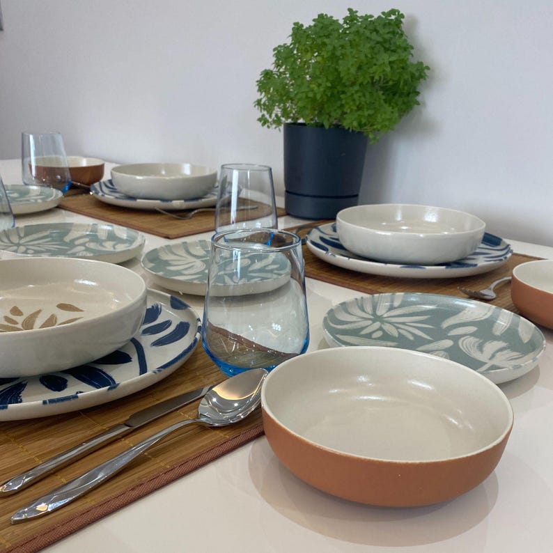 May include: A table setting featuring white and blue patterned plates, bowls, and clear glasses with blue rims. Silverware and small brown placemats are also visible. A potted plant adds a touch of greenery to the scene, creating a fresh and inviting dining atmosphere.