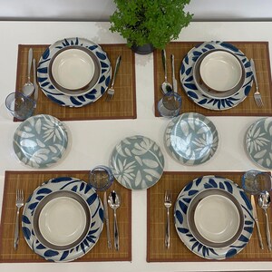 May include: A table setting for four featuring plates, bowls, and glasses. The plates have a blue and white floral design. Each place setting includes a bowl, a plate, a smaller plate, a glass, a fork, a knife, and a spoon. The table is set on bamboo placemats.