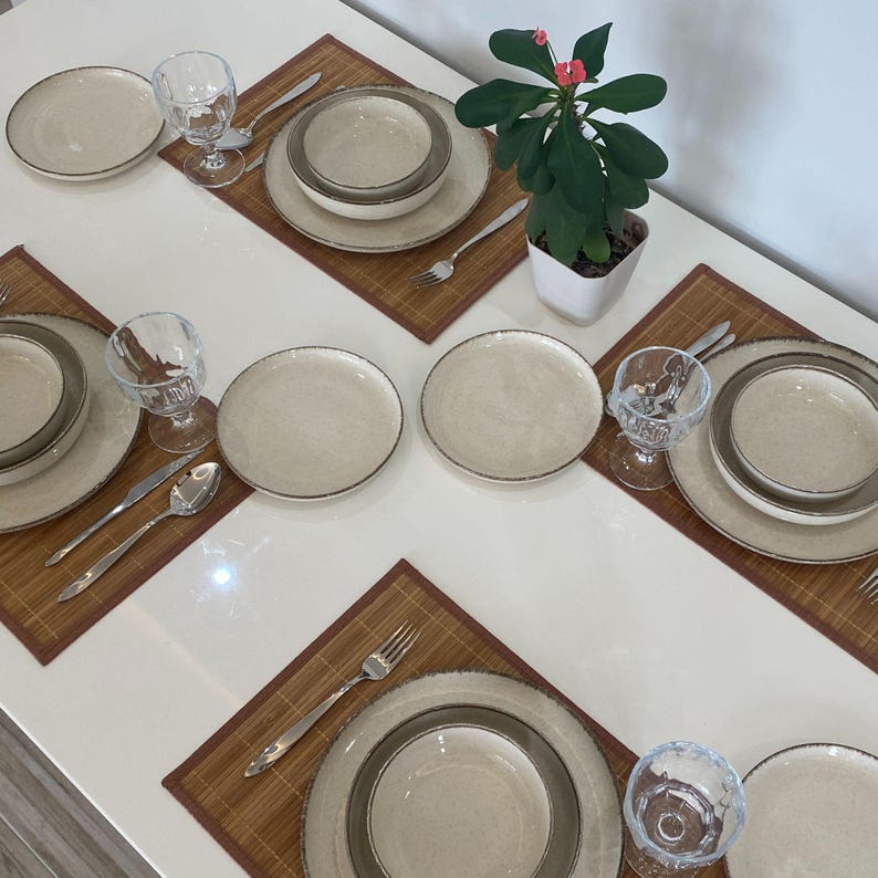 May include: A beige ceramic dinnerware set with a brown rim, including plates, bowls, and glasses, arranged on brown placemats. Silverware is placed next to each setting. A small potted plant with red flowers is in the background.