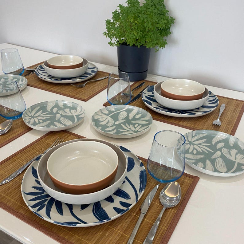 May include: A table setting featuring ceramic plates, bowls, and glasses. The plates have a white background with blue leaf designs. Bowls are brown and white. Clear glasses have blue rims. The setting includes silverware and bamboo placemats.