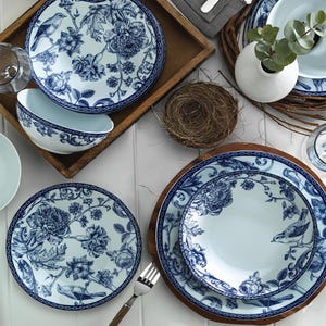Op de afbeelding: Een tafelsetting met blauw en wit bloemmotief servies. De set bevat borden, kommen en een kleine vaas met groen. Een houten dienblad en een nest maken deel uit van de decoratieve opstelling.