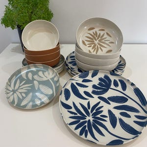 May include: A collection of ceramic dinnerware featuring floral designs. The set includes bowls and plates in various colors, including white, brown, and blue. The plates have a white background with blue floral patterns, while the bowls have a solid color.