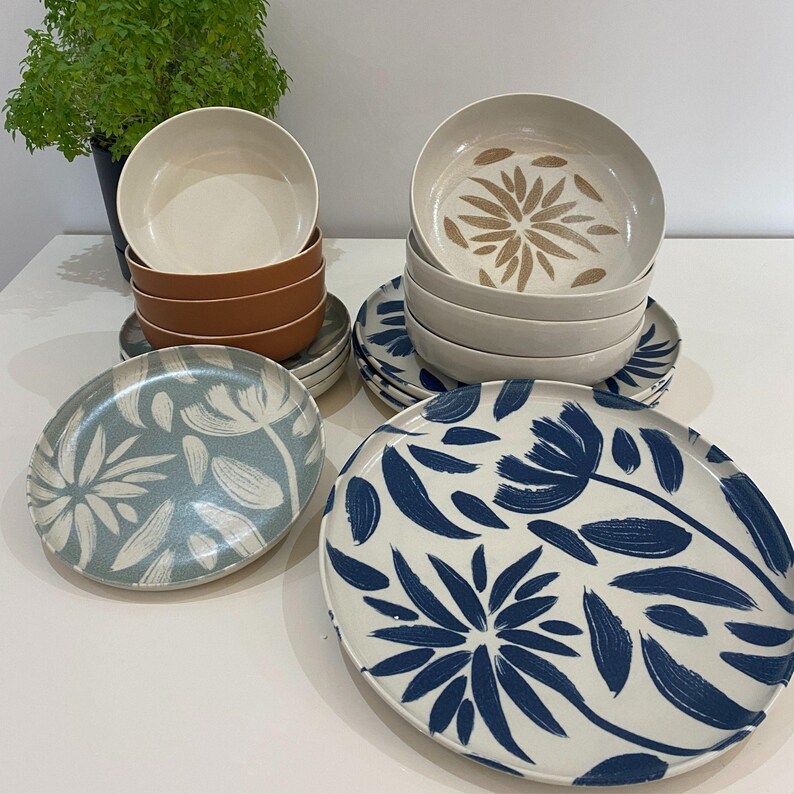 May include: A collection of ceramic dinnerware featuring floral designs. The set includes bowls and plates in various colors, including white, brown, and blue. The plates have a white background with blue floral patterns, while the bowls have a solid color.