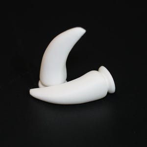 May include: Two white, curved, horn-shaped objects are displayed against a black background. One is upright, and the other is lying on its side. The objects have a smooth, glossy finish and a rounded base.