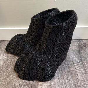 May include: A pair of black, woven-textured footwear resembling animal paws. The shoes have a sock-like design, with a rounded toe and a high ankle. The material appears to be a flexible, open weave.