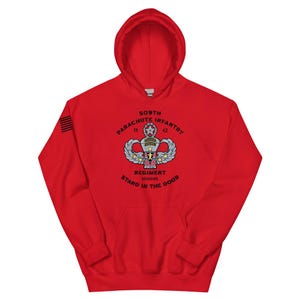 May include: A red hooded sweatshirt featuring a graphic of the 509th Parachute Infantry Regiment. The design includes the text "509th Parachute Infantry Regiment" and "Stand in the Door". The left sleeve has a small American flag.