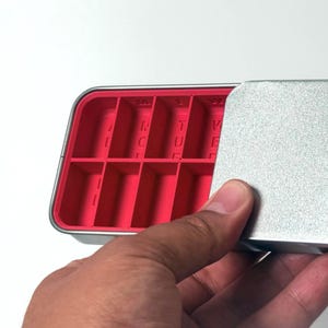 May include: A rectangular, silver-colored metal container with a bright pink interior divided into multiple compartments. The lid is partially open, revealing the compartments. The letters "R", "T", and "V" are visible inside the compartments.