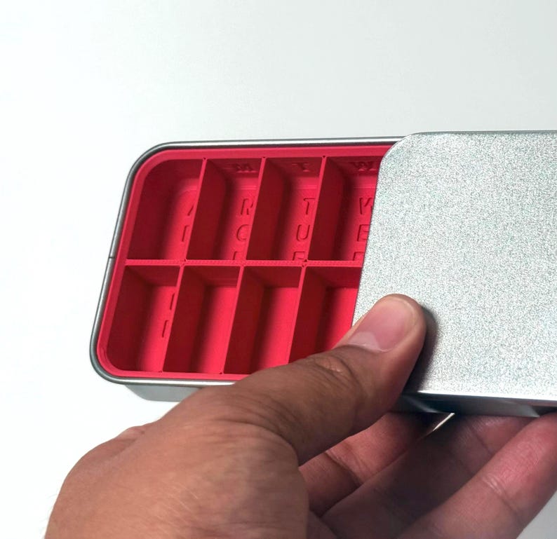 May include: A rectangular, silver-colored metal container with a bright pink interior divided into multiple compartments. The lid is partially open, revealing the compartments. The letters "R", "T", and "V" are visible inside the compartments.