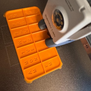 May include: An orange 3D-printed pill organizer with the days of the week and AM/PM labels. The organizer is rectangular with a grid of compartments and is being printed by a 3D printer. The text "Bambu Lab" is visible on the printer.