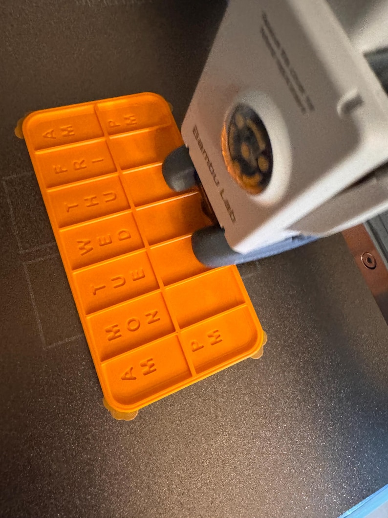 May include: An orange 3D-printed pill organizer with the days of the week and AM/PM labels. The organizer is rectangular with a grid of compartments and is being printed by a 3D printer. The text "Bambu Lab" is visible on the printer.