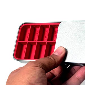 May include: A rectangular, silver metal container with a red interior divided into compartments. The lid is partially open, revealing the compartments. The container is being held by a hand.