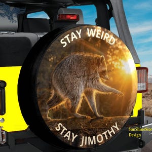 May include: A circular black spare tire cover featuring a graphic of a raccoon walking on a log at sunset. The text STAY WEIRD is printed in white at the top, and the text STAY JIMOTHY is printed in white at the bottom of the circular design.