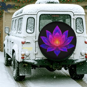May include: A white SUV covered in snow with a spare tire cover featuring a vibrant lotus flower design in shades of purple, pink, and yellow against a black background. The vehicle is parked in a snowy environment.