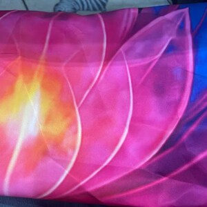 May include: A close-up of a fabric with a floral design. The image features a large, stylized flower with petals in shades of pink, orange, and yellow. The edges of the petals are outlined in white, and the background is a deep blue.