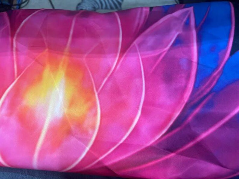 May include: A close-up of a fabric with a floral design. The image features a large, stylized flower with petals in shades of pink, orange, and yellow. The edges of the petals are outlined in white, and the background is a deep blue.