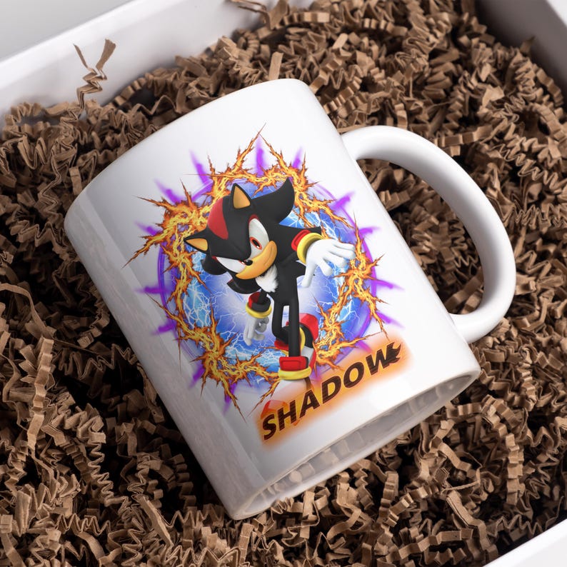 May include: White ceramic mug with a black and red cartoon character, Shadow the Hedgehog, in a blue and yellow energy field. The text "SHADOW" is printed in yellow on the mug.