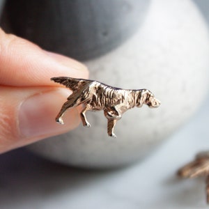 May include: A small, gold-colored dog figurine. The miniature sculpture depicts a dog in a walking pose, with detailed fur texture. The dog is held in a person's hand, with a blurred background.