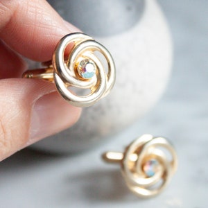 Könnte beinhalten: Goldfarbene Manschettenknöpfe mit einem wirbelnden, floralen Design. Jeder Manschettenknopf hat einen zentralen, irisierenden Kristall. Die Manschettenknöpfe werden vor einem neutralen Hintergrund präsentiert.