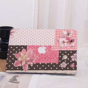 May include: A decorative laptop case featuring a patchwork design with floral and polka dot patterns in shades of pink, brown, and white. The case includes a white Apple logo and a lace trim along the bottom edge.
