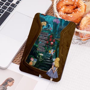May include: A brown phone case with a vibrant Alice in Wonderland illustration. The design includes the text "Wonderland" and signposts. The case is placed on a laptop, with a pretzel basket nearby.
