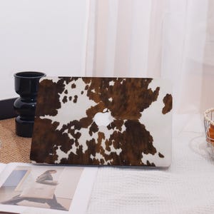 Puede incluir: Una funda para portátil con un estampado de piel de vaca marrón y blanco. La funda tiene un logotipo de Apple blanco en el centro. La funda está sobre una superficie blanca.