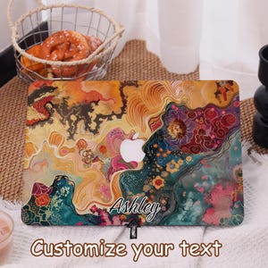 Op de afbeelding: Een laptophoesje met een kleurrijk abstract ontwerp met wervelingen van oranje, geel, teal en roze. Het Apple-logo is zichtbaar. De naam "Ashley" is cursief geschreven. De tekst "Customize your text" staat onderaan.