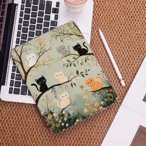 May include: A tablet case featuring a whimsical illustration of cats perched on tree branches. The design includes various colored cats, including white, gray, black, and orange, set against a soft green and floral background.