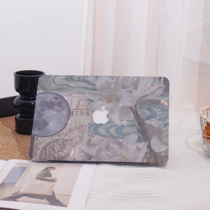 May include: A laptop case featuring a floral design with a full moon, turtle, and the word "MINA". The case has a white Apple logo and a light blue and white color scheme. The case is on a white surface.