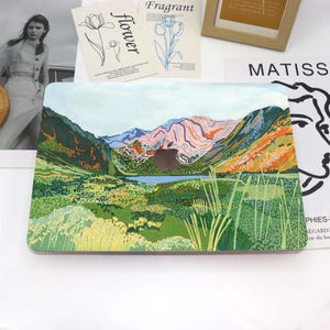 May include: A laptop skin with a colorful illustration of a mountain range and a lake. The mountains are painted in shades of pink, green, and blue, with a small lake in the foreground. The skin is designed to fit a standard laptop.