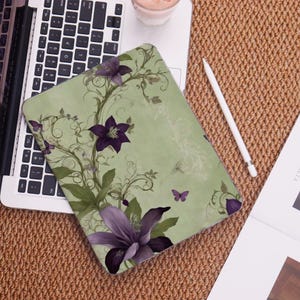 May include: A light green laptop case featuring a floral design with purple flowers, green leaves, and butterflies. The case is resting on a laptop keyboard and a woven brown surface. A white stylus and a glass of pink liquid are also visible.