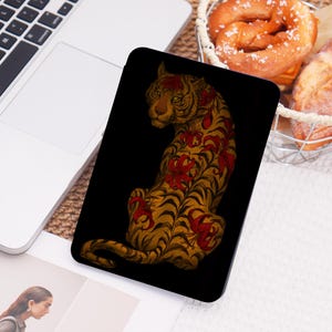 May include: A black tablet case featuring a detailed illustration of a tiger. The tiger is depicted in shades of gold and brown with red floral accents. The design is set against a solid black background.