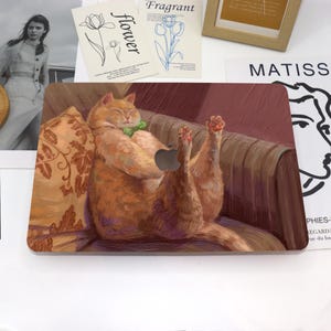 Könnte beinhalten: Eine Laptop-Haut mit einem Gemälde einer orangefarbenen Tabby-Katze, die auf einem Sofa mit hoch erhobenen Pfoten liegt. Die Katze trägt eine grüne Fliege und ist von einem floralen Muster umgeben.