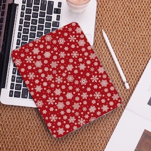 May include: A red tablet case with a white snowflake pattern. The case is on top of a laptop keyboard. A white stylus and a cup of coffee are also visible on the surface.