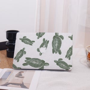 Può includere: Una custodia bianca per laptop con un motivo di tartarughe marine verdi. Le tartarughe sono raffigurate in varie pose, creando un design ispirato alla natura. La custodia è progettata per adattarsi a un laptop, offrendo protezione e un'estetica unica.