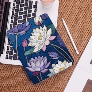 Könnte beinhalten: Eine blaue Tablet-Hülle mit einem floralen Design aus weißen und lila Lotusblumen. Die Hülle liegt auf einer Laptop-Tastatur, mit einem weißen Stift und einem Getränk im Hintergrund. Die Lotusblumen haben grüne Blätter und sind vor einem dunkelblauen Hintergrund.