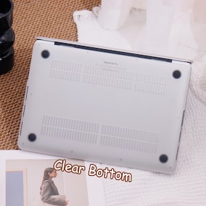 May include: A clear bottom laptop case with the text "Clear Bottom" in a brown font. The case is designed for a MacBook Pro and has ventilation slots. The case is light gray and has four black rubber feet.